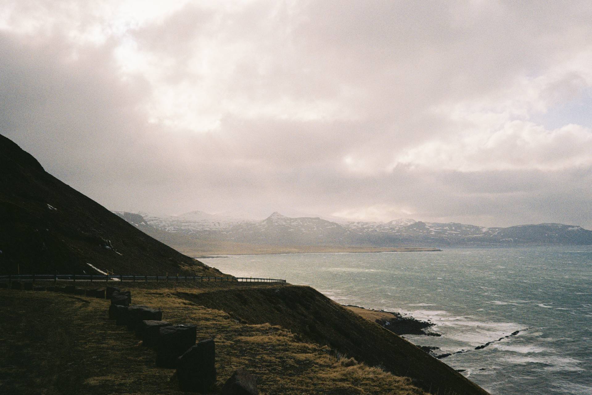 A landscape photograph of the sea, with a road in the foreground and some mountains in the background